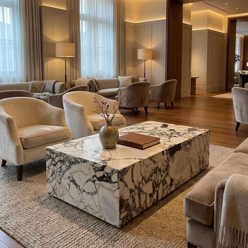 A modern rectangular coffee table with a white marble-look surface featuring dramatic black and gray veining, styled in a luxurious, warm-toned living room with cream armchairs and a woven rug.