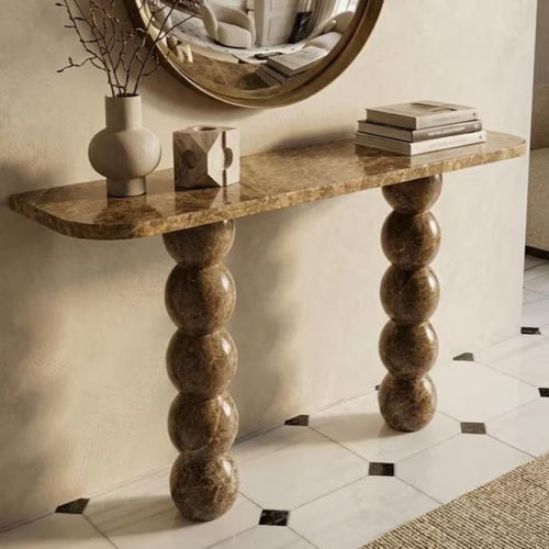 A console table with a warm brown marble-look top and sculptural stacked-bulb legs, styled in an interior setting with a vase, decorative blocks, books, and a round mirror.