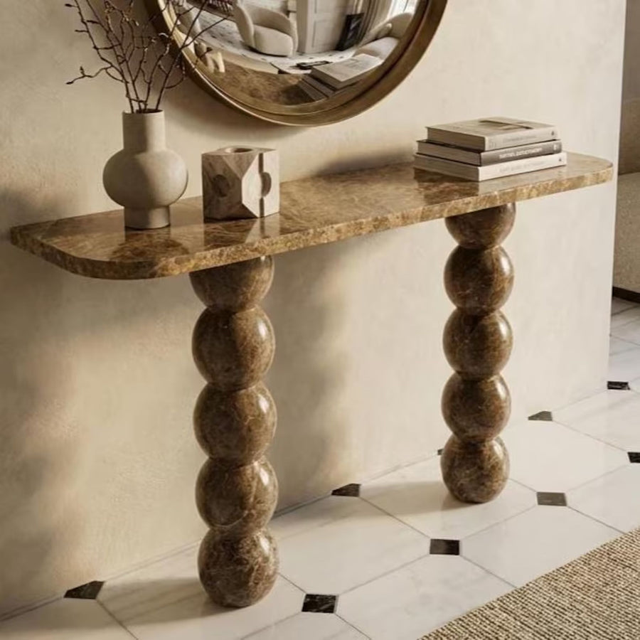 A console table with a warm brown marble-look top and sculptural stacked-bulb legs, styled in an interior setting with a vase, decorative blocks, books, and a round mirror.
