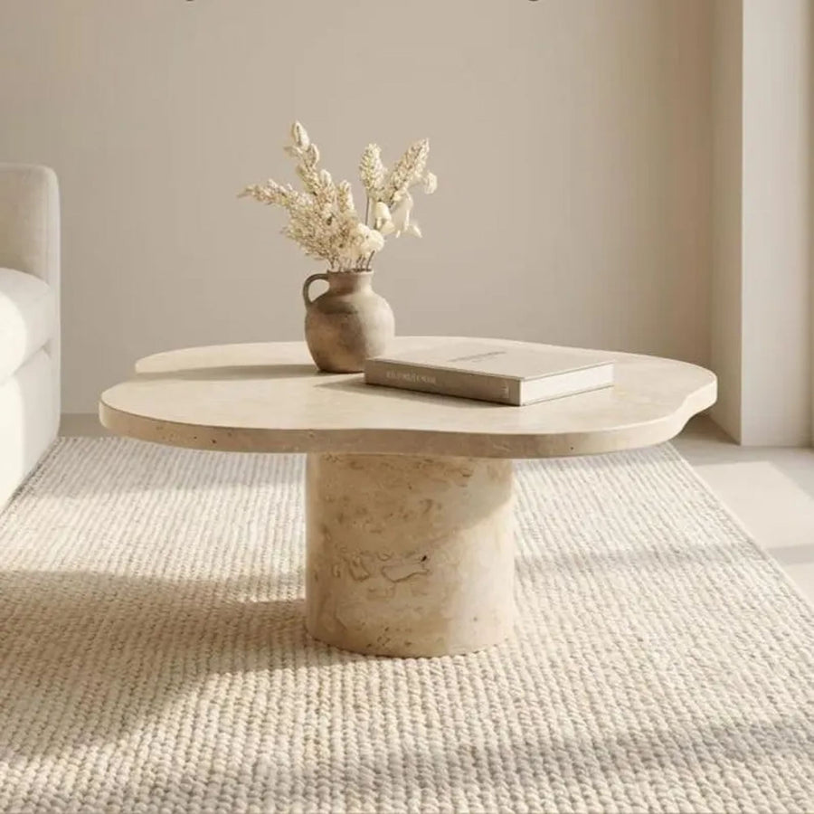 A round stone coffee table with a textured cylindrical base, styled with a small vase of dried flowers and a book on a woven rug in a softly lit room.