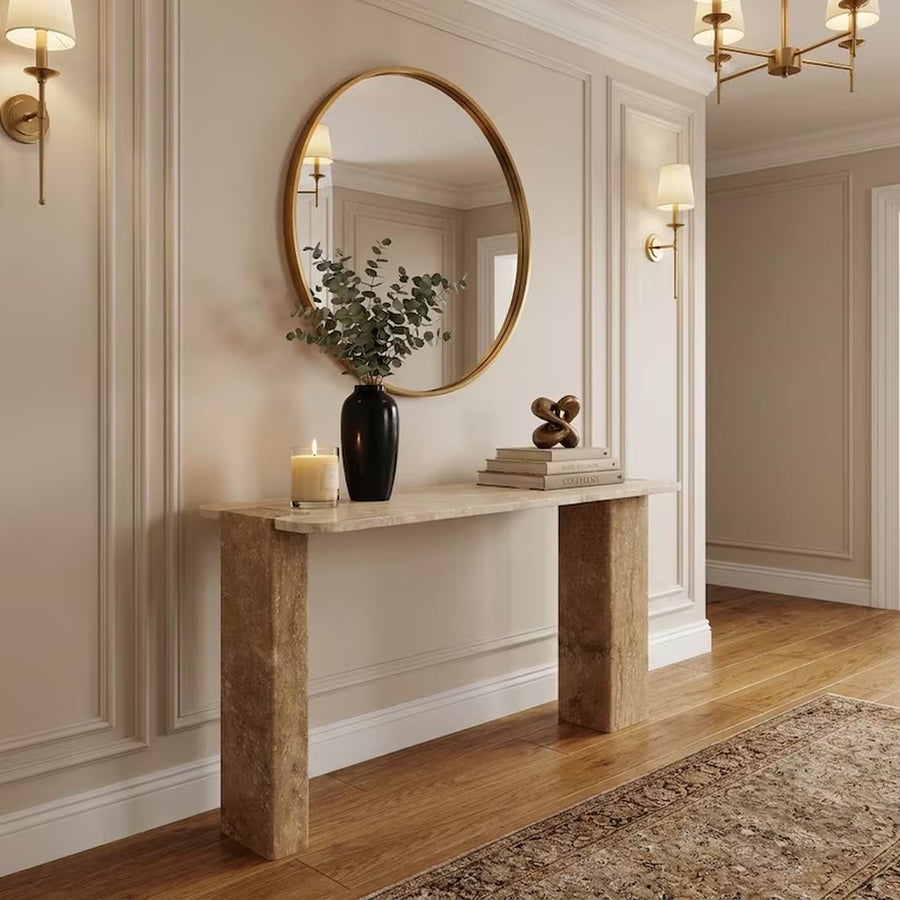 A rectangular console table with a light beige stone top and blocky stone legs, styled in a classic entryway with a gold-framed oval mirror, wall sconces, and a wooden floor.