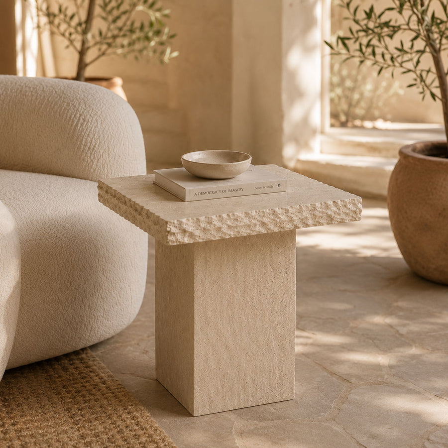 A square side table with a textured stone top and a solid block pedestal base in a warm beige tone, styled in a sunlit room next to a cream-colored textured sofa.