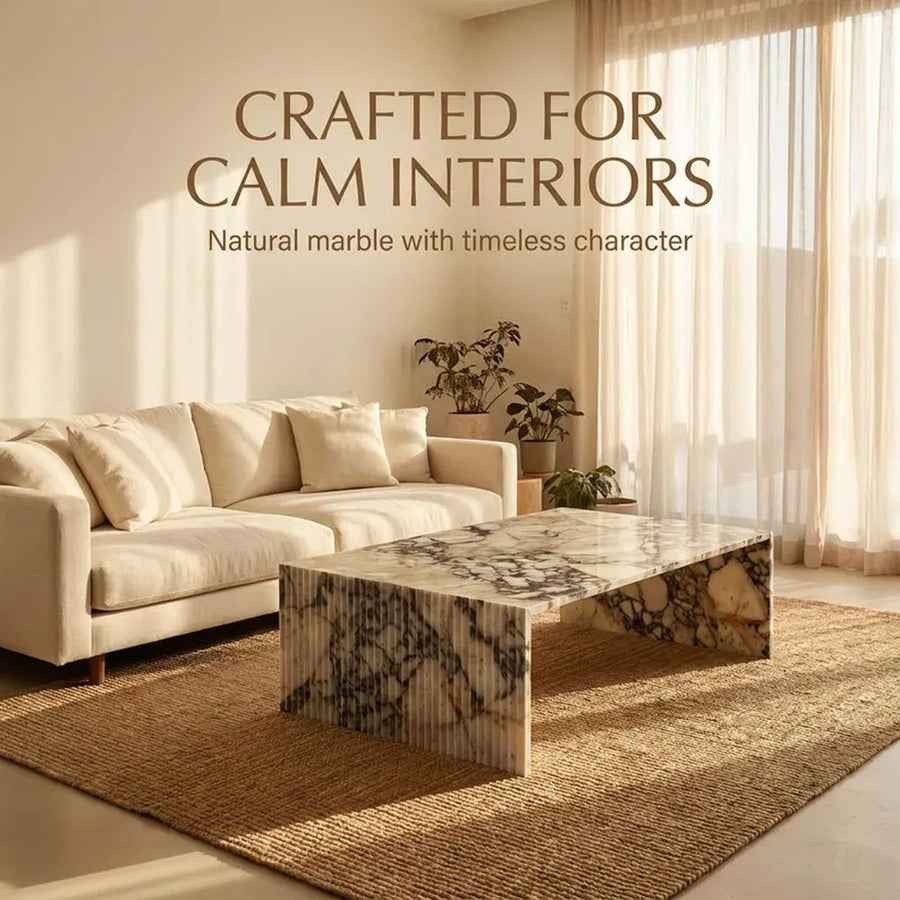 A rectangular coffee table with a natural marble top and fluted base, styled in a warm, minimalist living room next to a cream sofa and a woven rug. Text overlay reads 'CRAFTED FOR CALM INTERIORS' and 'Natural marble with timeless character'.