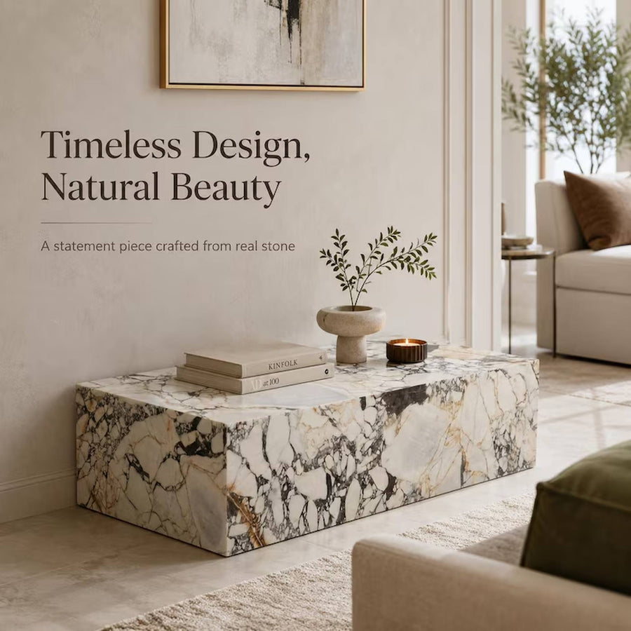 A rectangular stone coffee table with a white marble-look surface and black, gray, and warm gold veining, styled in a modern living room setting with books, a small plant, and a candle. Text on the wall reads 'Timeless Design, Natural Beauty' and 'A statement piece crafted from real stone'.