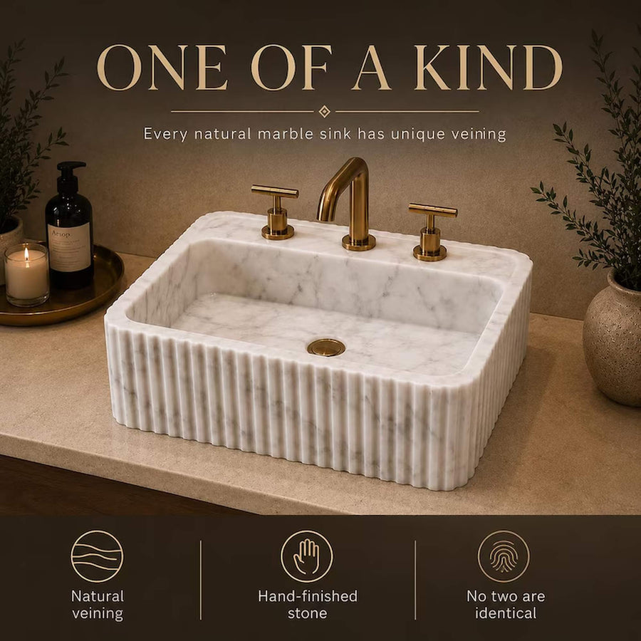 A rectangular white marble sink with fluted sides, a single basin, and a matching brass faucet set, shown on a countertop with decorative plants and bottles. Text at the top reads 'ONE OF A KIND' and the bottom banner highlights natural veining, hand-finished stone quality, and uniqueness.