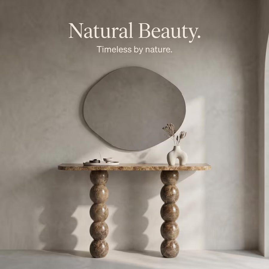 A styled interior scene with a large organic-shaped wall mirror above a stone console table with bulb-shaped legs. A small white vase with dried stems sits on the table, and the wall has the text 'Natural Beauty.