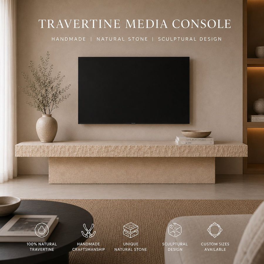 A travertine media console in a modern living room setting, featuring a textured natural stone console table with a matching floating shelf and a built-in base. A black flat-screen TV is mounted on the beige wall above, with a ceramic vase and books on the shelf.