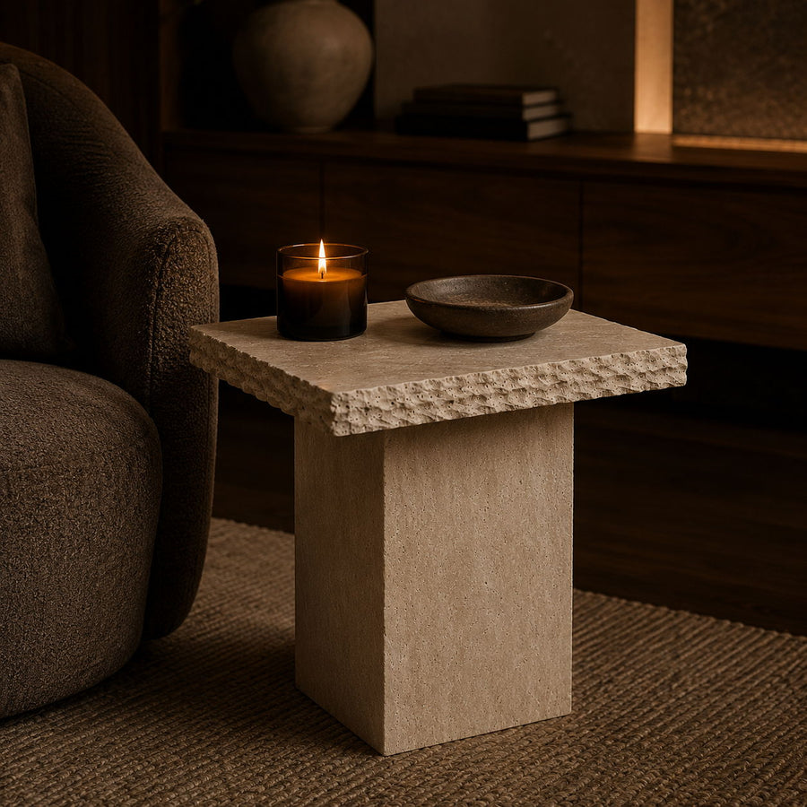 A small rectangular stone side table with a textured top, styled in a cozy living room next to a dark armchair with a lit candle and a bowl on top.