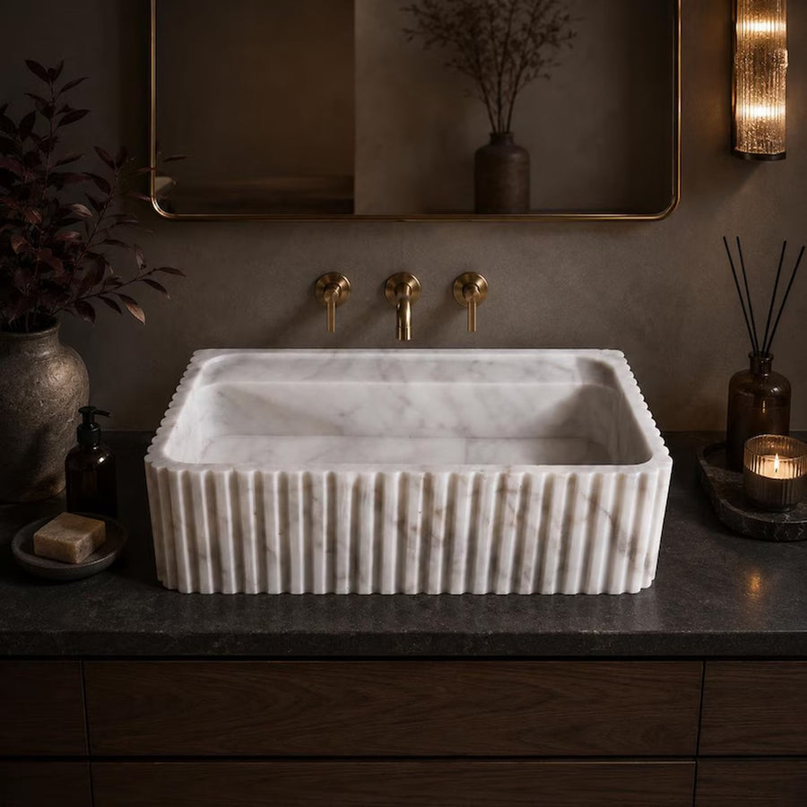 A rectangular white marble vessel sink with a fluted front edge, set into a dark countertop with a gold faucet and brass fixtures in a styled bathroom setting.