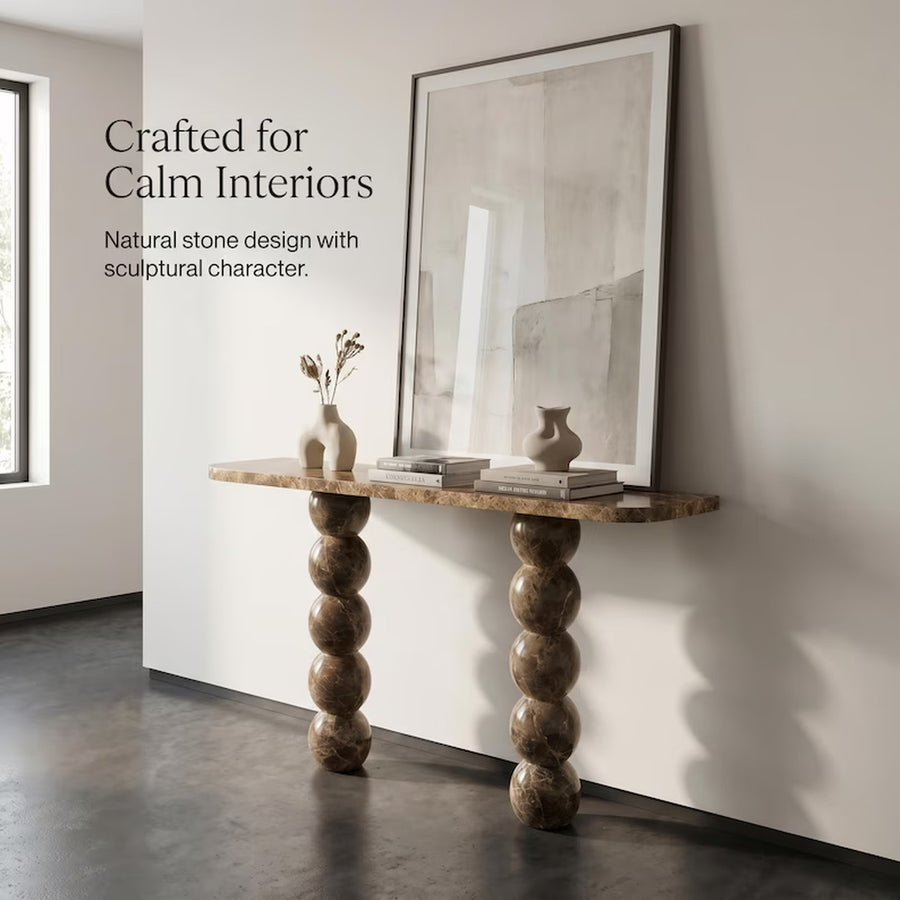 A console table with a natural stone top and two sculptural, stacked-bubble legs in a warm brown tone, styled with vases and books against a white wall in a minimalist interior. Text on the left reads: 'Crafted for Calm Interiors' and 'Natural stone design with sculptural character.