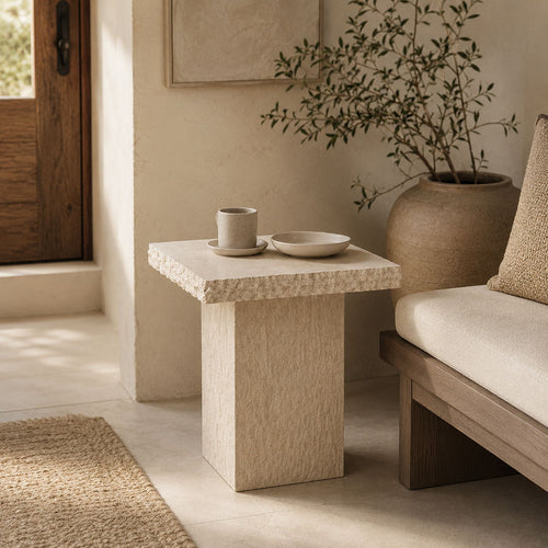 A square stone end table in a warm beige textured finish, styled in a minimalist interior next to a wooden bench with a cream cushion. On the table are a small ceramic cup and saucer and a shallow bowl.
