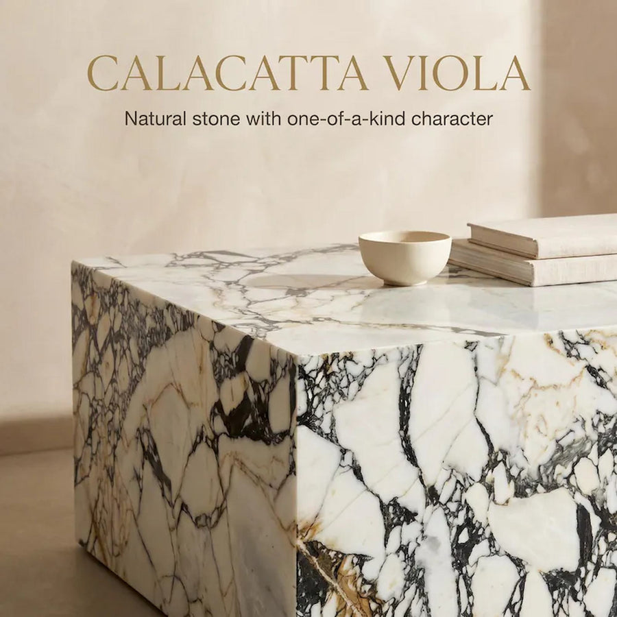 A polished Calacatta Viola marble block or side table with dramatic black, white, and gold veining, styled with a small cream bowl and books on top. Text at the top reads 'CALACATTA VIOLA' and 'Natural stone with one-of-a-kind character'.