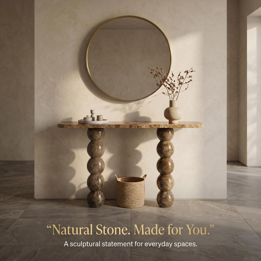 A sculptural console table with a natural stone top and twisted, bulb-like stone legs stands against a textured beige wall in a styled interior. A round gold-framed mirror hangs above, with a small vase of dried branches on the right and a woven basket beneath the table.