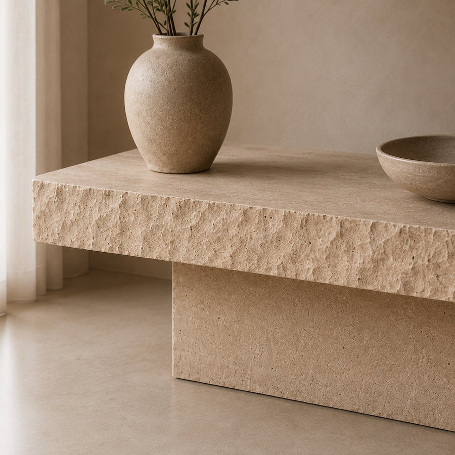 A modern low coffee table made of textured beige stone, styled with a matching ceramic vase and a ceramic bowl in a minimalist room.