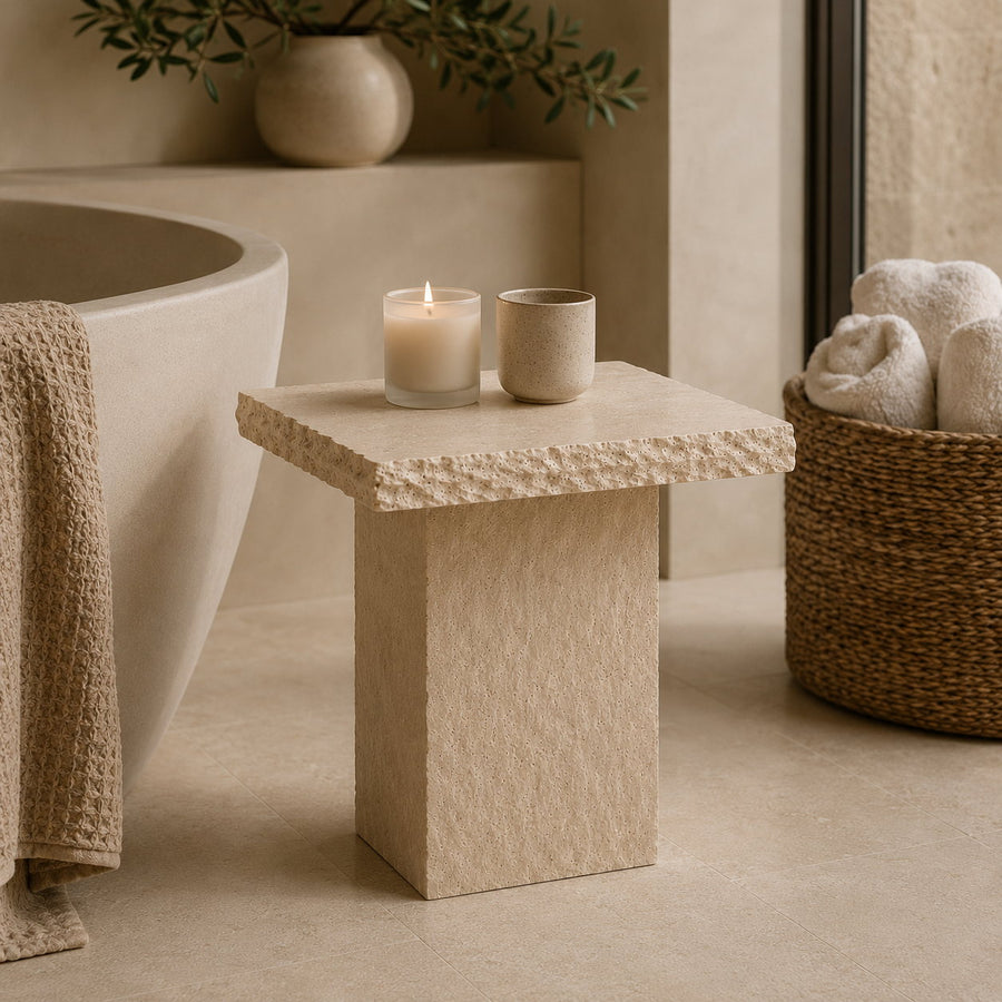 A small square stone side table with a textured top stands in a warm neutral bathroom setting beside a bathtub, with two candles on top and a woven basket of towels nearby.