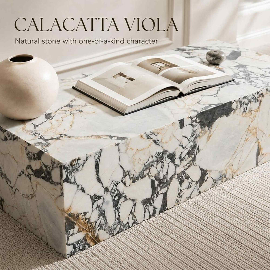 A rectangular stone bench or coffee table made of Calacatta Viola marble, styled with an open book, a small ceramic vase, and a stone orb in a bright interior setting.