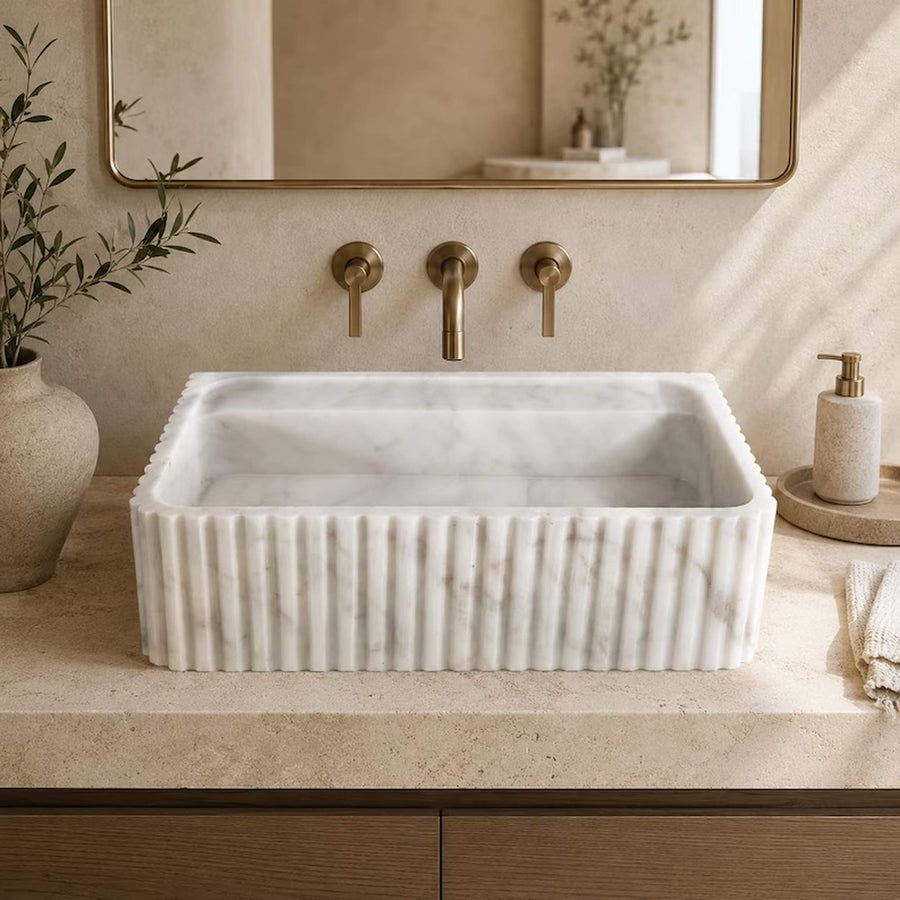 A rectangular white marble vessel sink with a fluted front edge, set into a beige stone countertop with a matching beige soap dispenser, a vase with greenery, and a brass faucet set beneath a mirror.
