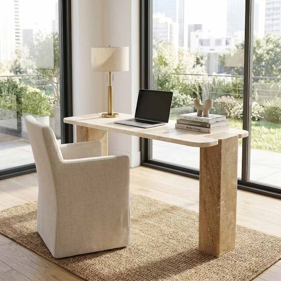 A modern home office setup featuring a light beige stone desk with a matching upholstered armchair, styled with a lamp, laptop, and books. The scene is set in a bright room with large windows overlooking a green outdoor patio.