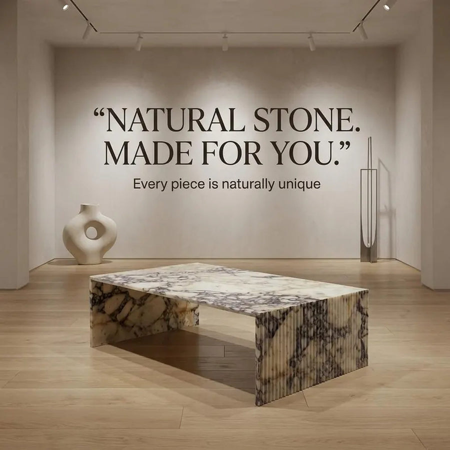 A rectangular coffee table with a polished natural stone top and ribbed fluted sides, placed in a minimalist modern room with light wood floors and a beige wall featuring the quote 'NATURAL STONE. MADE FOR YOU.