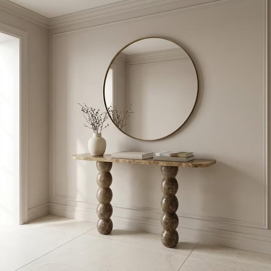 A round wall mirror with a thin gold-toned frame hangs above a console table with a stone top and sculptural stacked legs. The room has warm beige walls, white trim, and a light floor, with a small vase and books styled on the table.