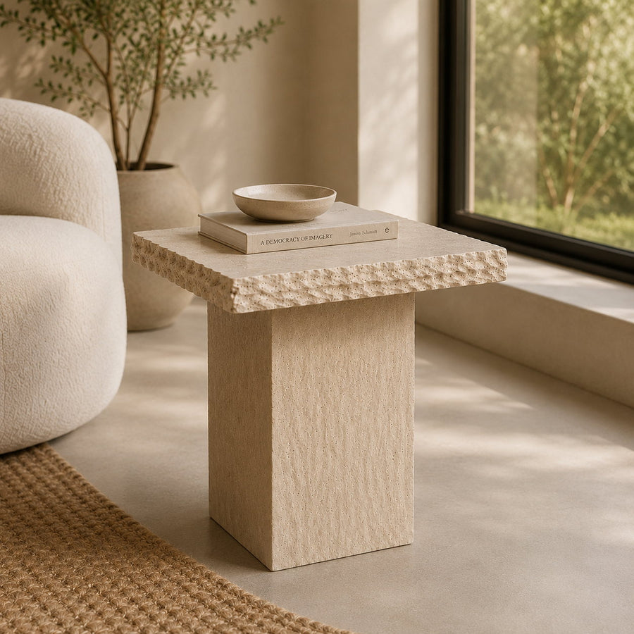 A light beige textured stone side table with a square top and block pedestal base, styled in a minimalist living room next to a white armchair and a window.