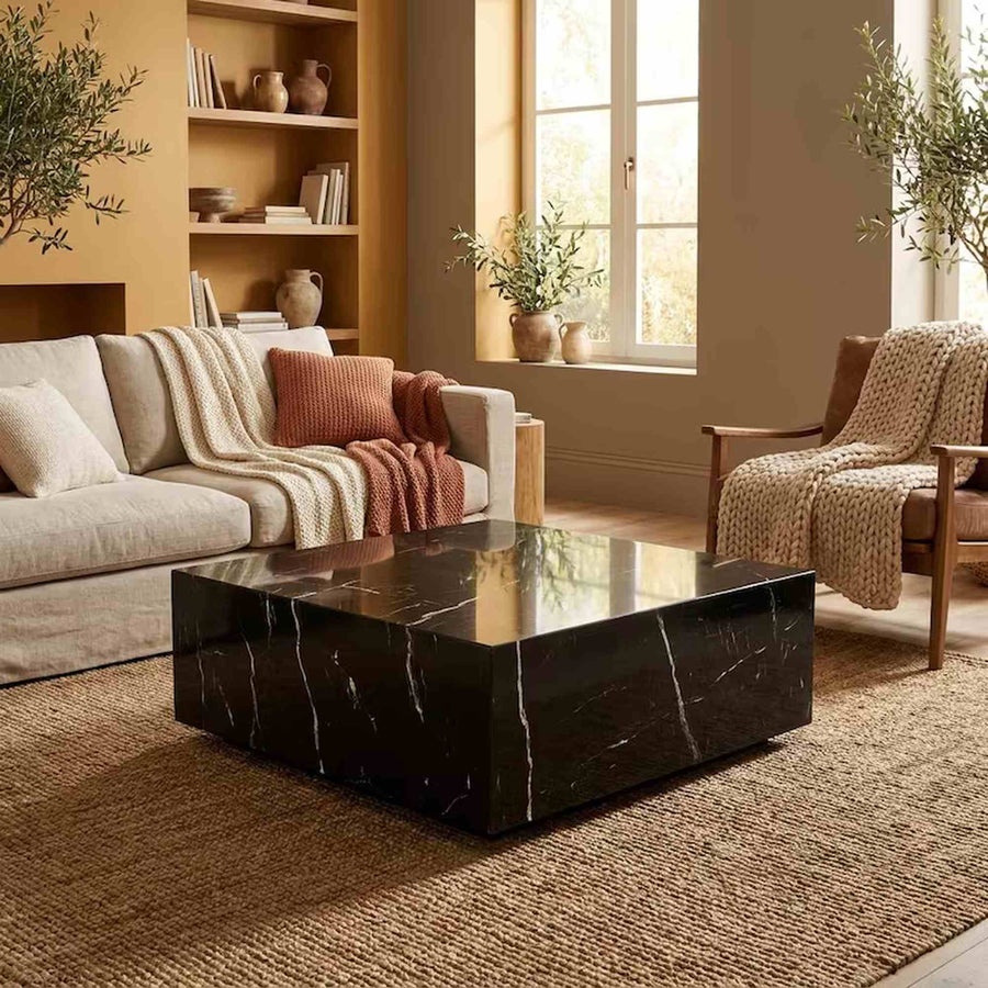 A modern living room featuring a glossy black marble coffee table with white veining, set on a woven jute rug. The room includes a beige sofa with neutral-toned cushions and throw blankets, a wooden armchair with a knit throw, a built-in wooden bookshelf, and warm natural light coming from a window.