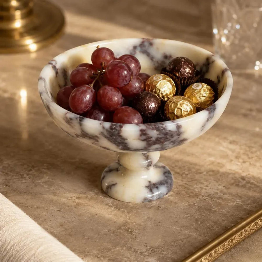 A marble pedestal bowl filled with red grapes and wrapped chocolates, set on a beige stone surface with a blurred warm-toned background.