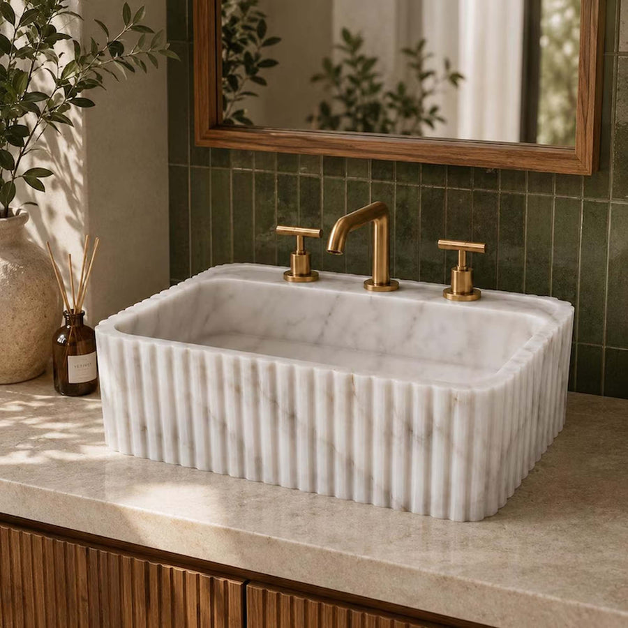 A white marble vessel sink with a fluted front edge, installed in a beige stone countertop. It is paired with a brushed brass faucet and matching handles, set against a dark green tiled backsplash and a wooden-framed mirror.