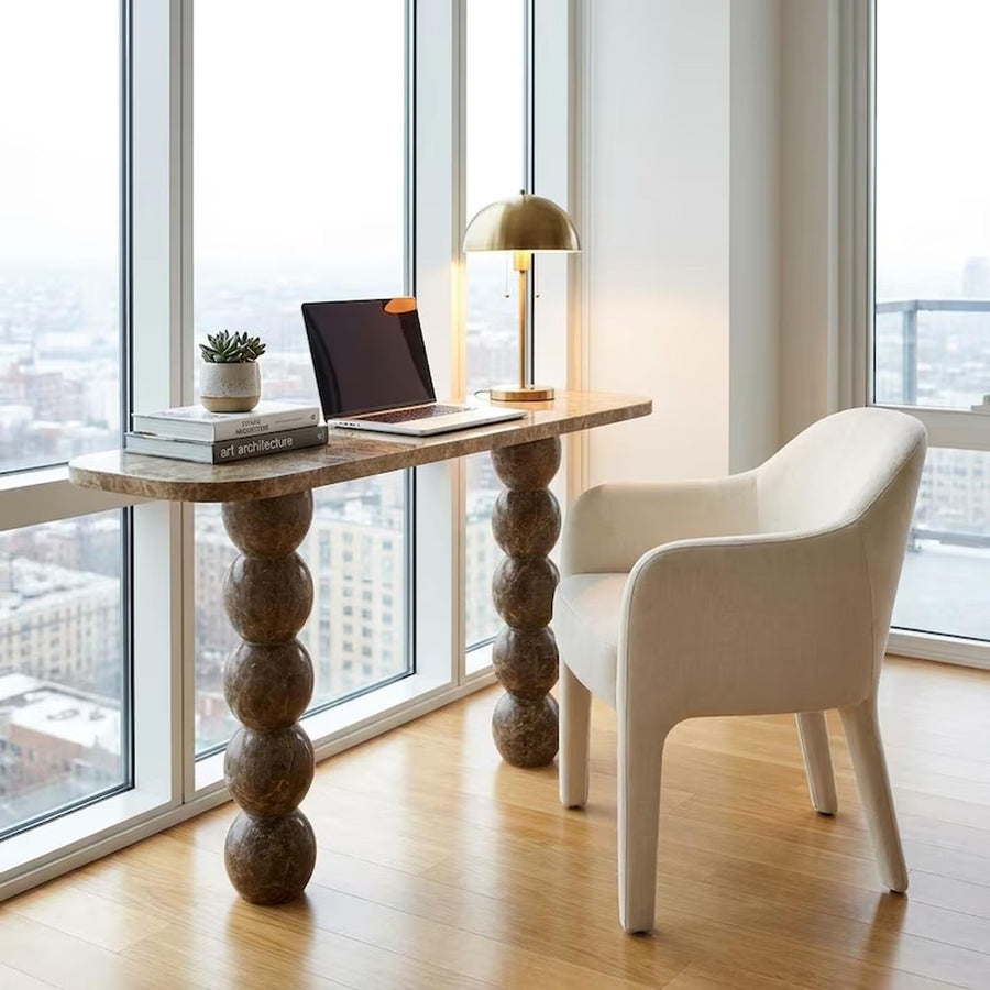 A modern home office setup featuring a long marble-top desk with chunky stacked ball legs, paired with a cream upholstered armchair and a brass table lamp, set against large windows overlooking a city skyline.
