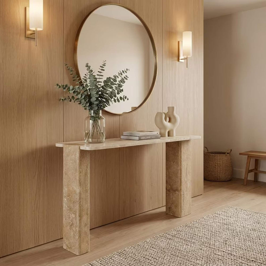 A modern console table with a light stone top and textured stone legs, styled with a glass vase of eucalyptus, books, and decorative objects, set against a light wood-paneled wall with a large round mirror and wall sconces.