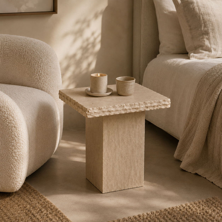 A small square side table with a textured beige stone-like top and block pedestal base, styled in a softly lit bedroom next to a cream boucle chair and bed.