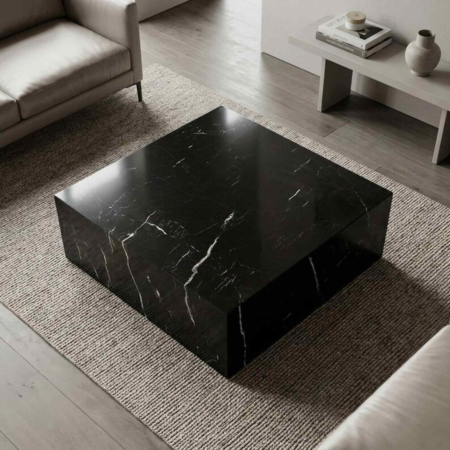 A square black marble coffee table with white veining, set on a textured beige rug in a modern living room.