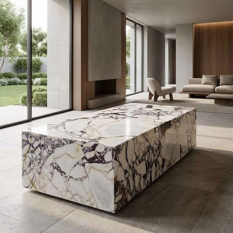 A rectangular marble coffee table with a polished cream, white, and brown veined surface sits in a modern, minimalist living room with large windows and a fireplace.