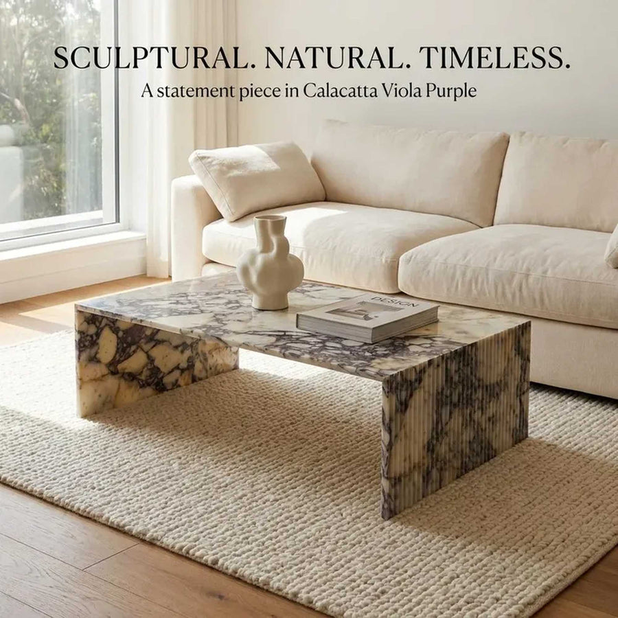 A sculptural rectangular coffee table made of Calacatta Viola Purple marble, styled in a bright living room with a cream sofa and a textured rug. Text overlay at the top reads 'SCULPTURAL.