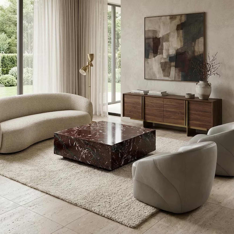 A modern living room featuring a curved beige sofa, two light gray armchairs, a dark reddish-brown marble coffee table, a wooden sideboard with gold accents, and a large abstract painting on the wall.