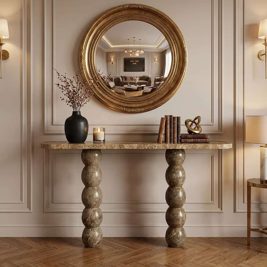 An ornate round gold-framed mirror hangs on a beige paneled wall above a marble console table with sculpted stone legs. The table is styled with a black vase, candles, books, and decorative objects, with a herringbone wood floor below.