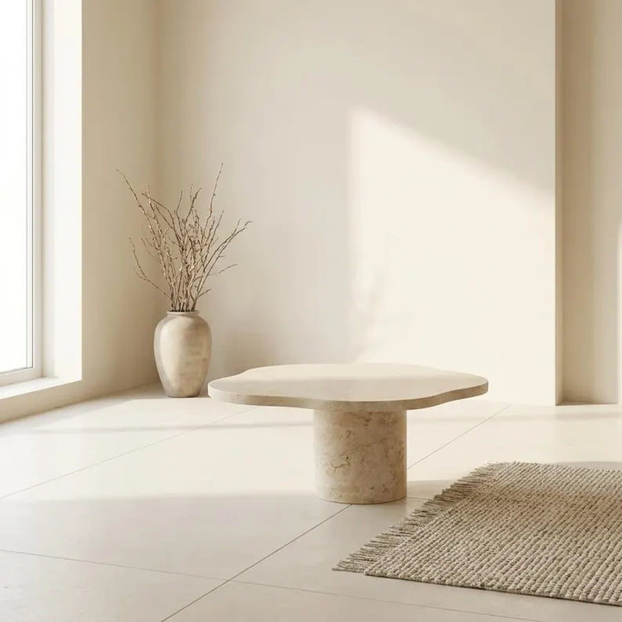 A modern low coffee table made of natural stone with a rounded organic top and cylindrical base, styled in a minimalist beige room with a woven rug, a vase of dried branches, and soft sunlight.