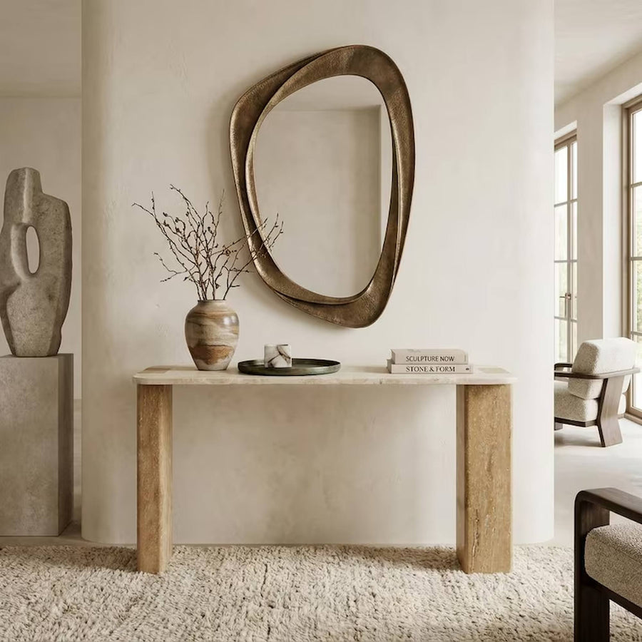 An abstract organic-shaped wall mirror with a distressed bronze finish hangs on a textured cream plaster wall above a light wood console table in a modern, minimalist living space.