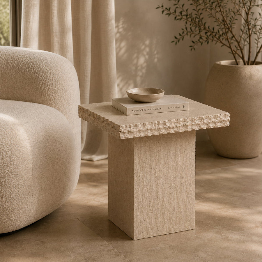 A square side table with a textured beige stone-like finish and a decorative beaded edge, styled in a warm, modern living room next to a curved cream sofa.