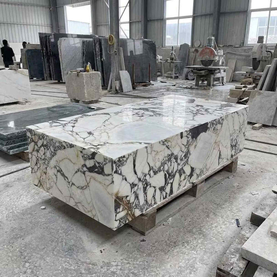 A large rectangular slab of polished white marble with dramatic black, gray, and warm gold veining, displayed on wooden pallets inside a stone processing factory.