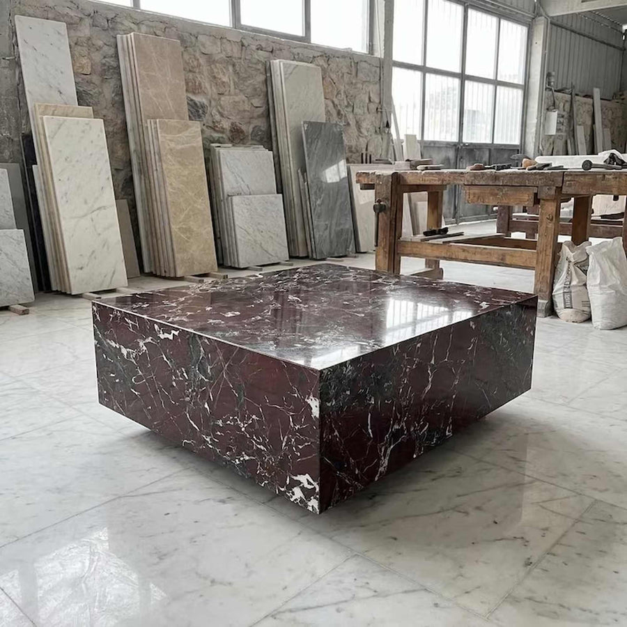 A square marble coffee table with a deep burgundy red marble pattern and white veining, set in a showroom with other marble slabs and a wooden workbench in the background.