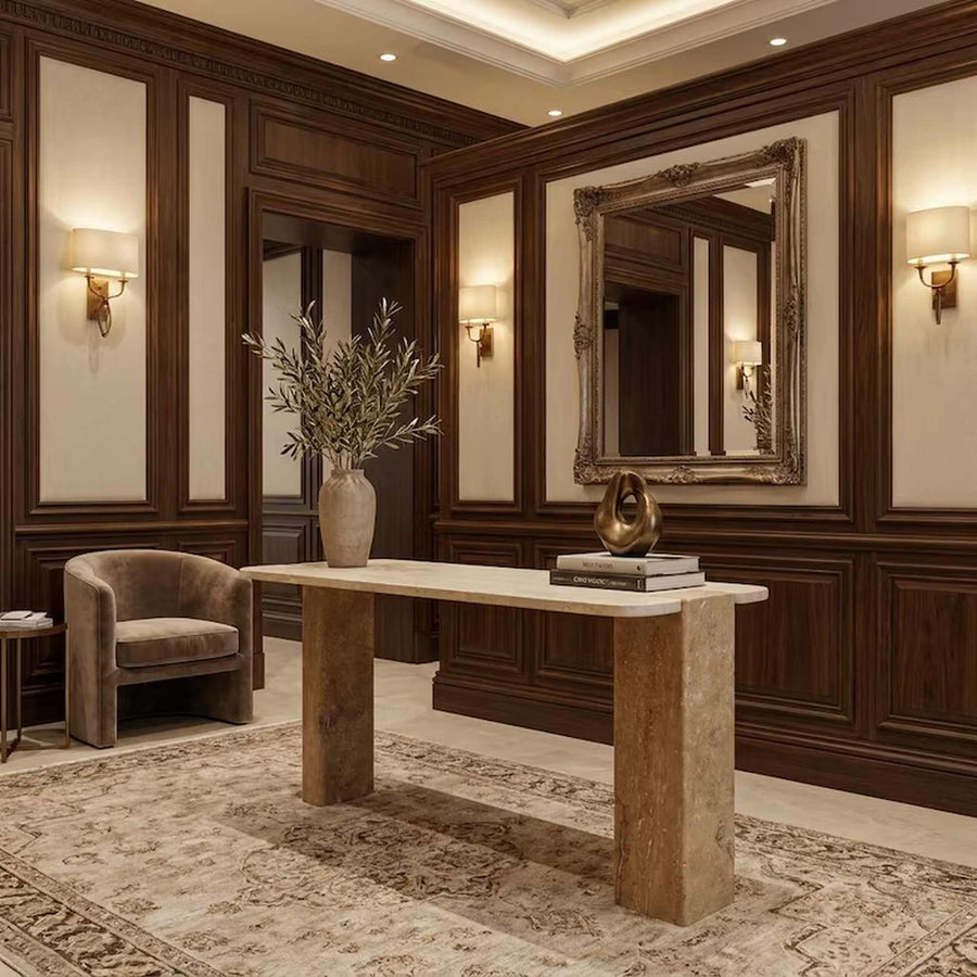 A luxury entryway featuring a large rectangular stone console table with a beige marble top, styled with a vase of olive branches and a small sculpture. The room has dark wood wainscoting, a large ornate gold-framed mirror, wall sconces, and a patterned area rug.