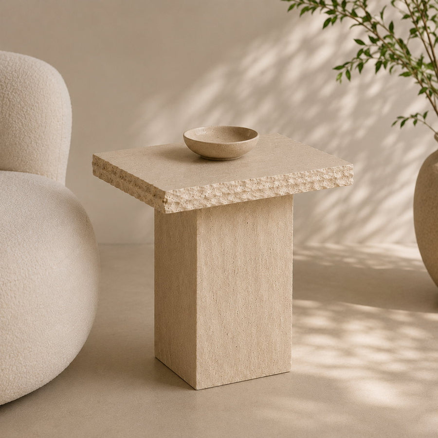 A minimalist beige stone side table with a textured flat top and block pedestal base, styled in a warm neutral room with a small matching bowl on top.