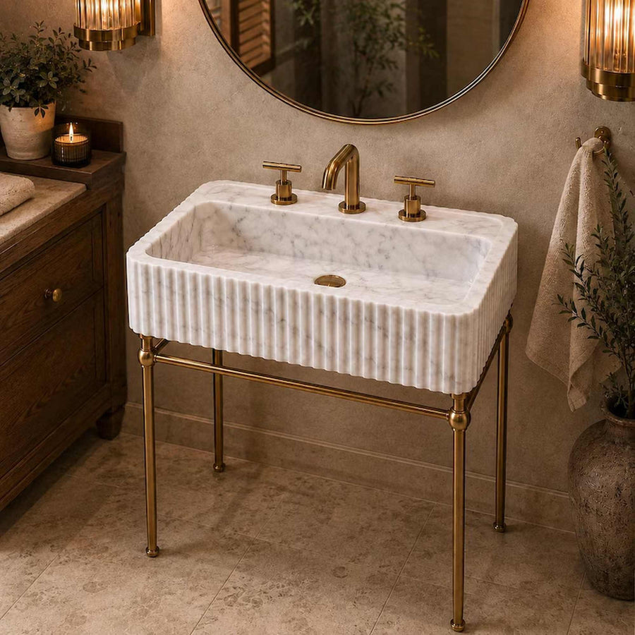 A white marble washbasin with a fluted edge and a brass stand, set in a warm, rustic bathroom with a wooden cabinet, round mirror, and warm lighting.