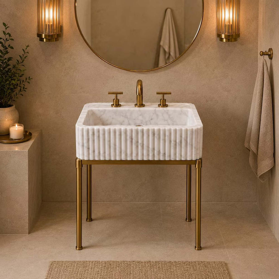 A modern bathroom sink with a white marble basin and a ribbed brass stand, set against a textured beige wall with a round mirror, wall sconces, and a hanging towel.