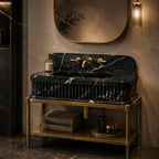 A luxury bathroom vanity featuring a black marble sink with white veining and a ribbed front, mounted on a gold metal stand. The setup includes a gold faucet, matching gold wall-mounted mirror, and warm ambient lighting against a textured beige wall.
