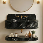 A black marble wall-mounted sink with white veining, fluted edges, and gold-tone faucet hardware, shown installed on a beige wall with a round mirror above and a floating shelf below holding rolled towels, a candle, bottles, and a small plant.