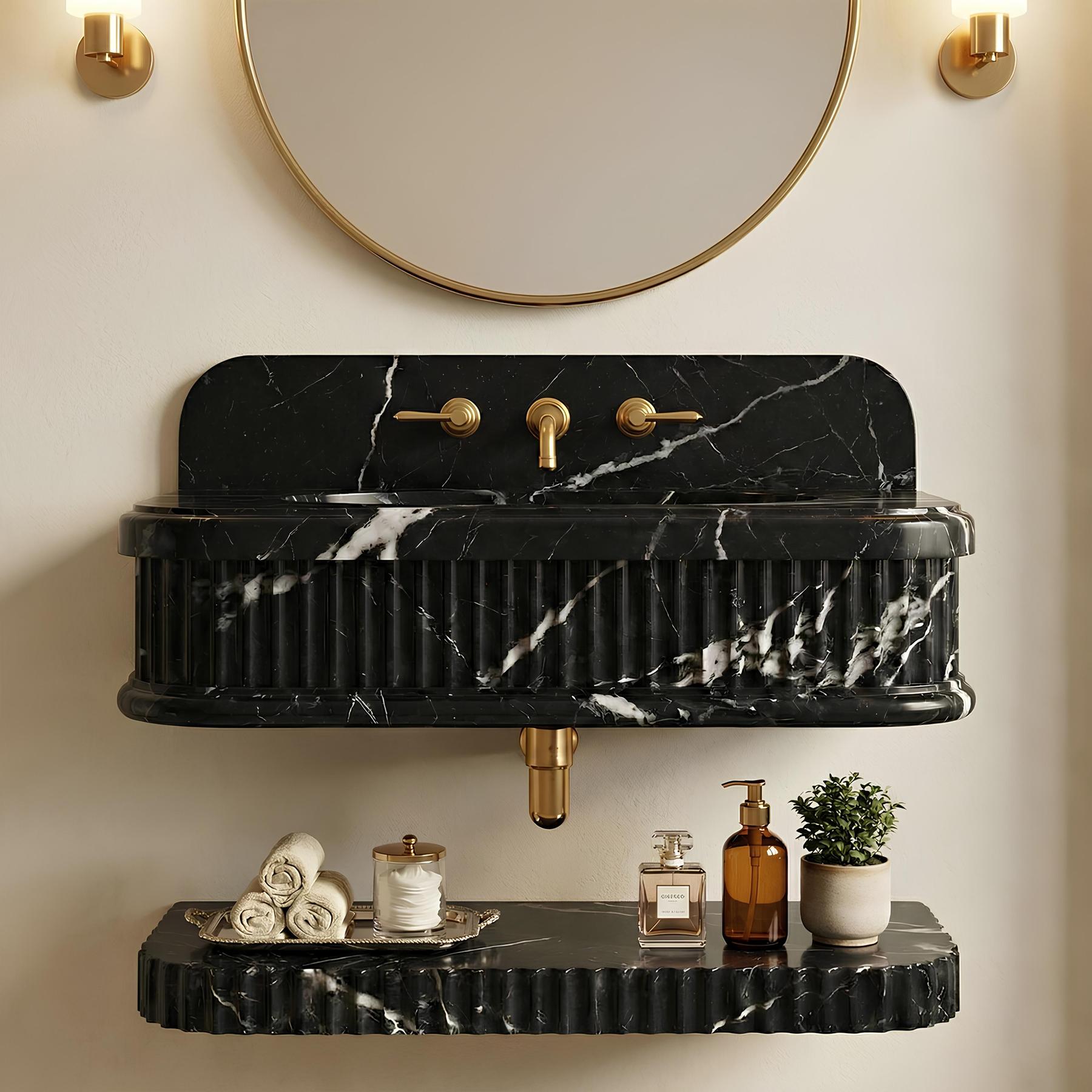 A black marble wall-mounted sink with white veining, fluted edges, and gold-tone faucet hardware, shown installed on a beige wall with a round mirror above and a floating shelf below holding rolled towels, a candle, bottles, and a small plant.