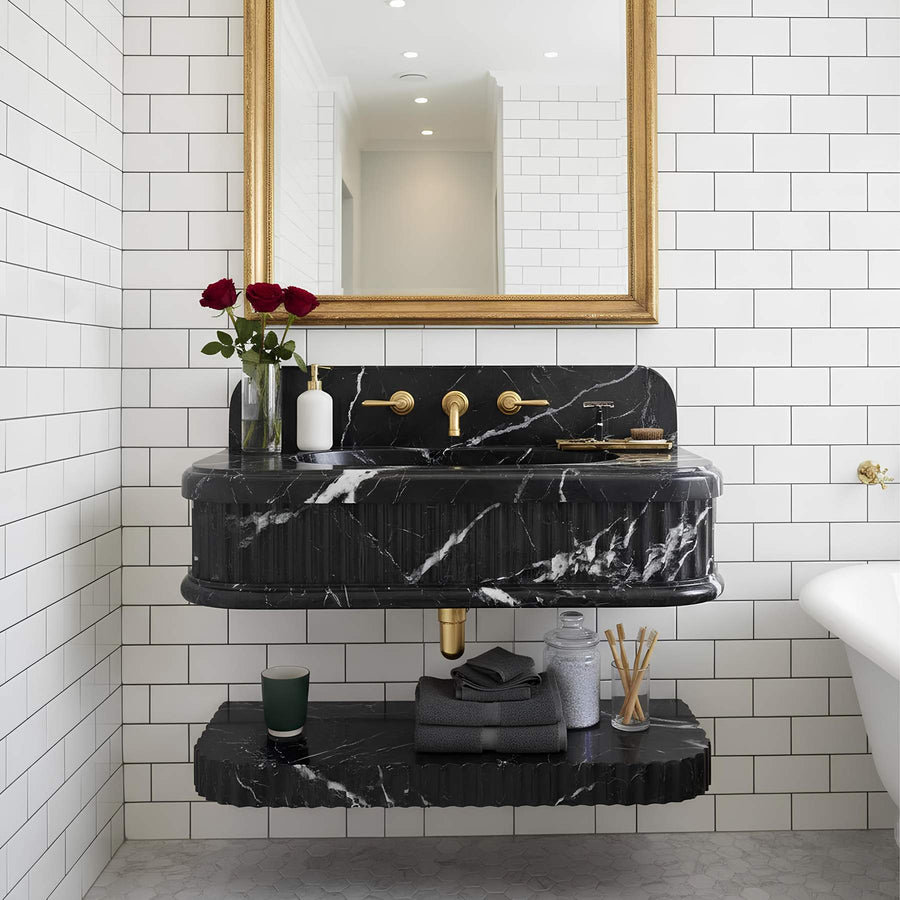 A modern bathroom vanity featuring a black marble sink and floating shelf with gold hardware, set against white subway tile walls under a gold-framed mirror.