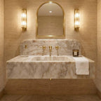 A luxury bathroom vanity featuring a floating marble sink with a white and beige veined pattern, gold fixtures, a matching marble backsplash, a gold-framed arched mirror, and two illuminated wall sconces. A folded white towel with a gold monogram is draped over the right side of the counter.